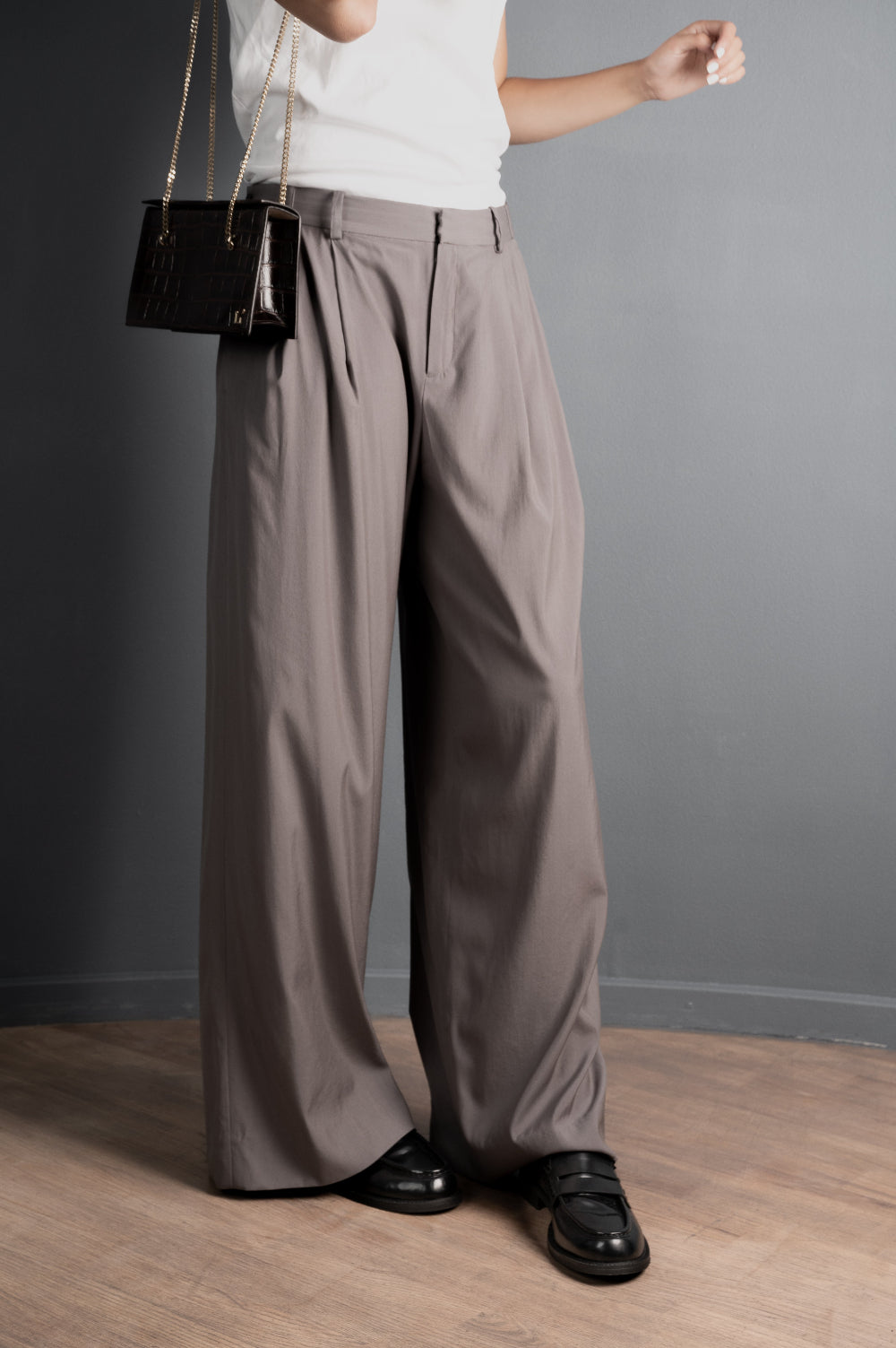 Person wearing brown wide-leg pants and a white top, standing against a gray wall.