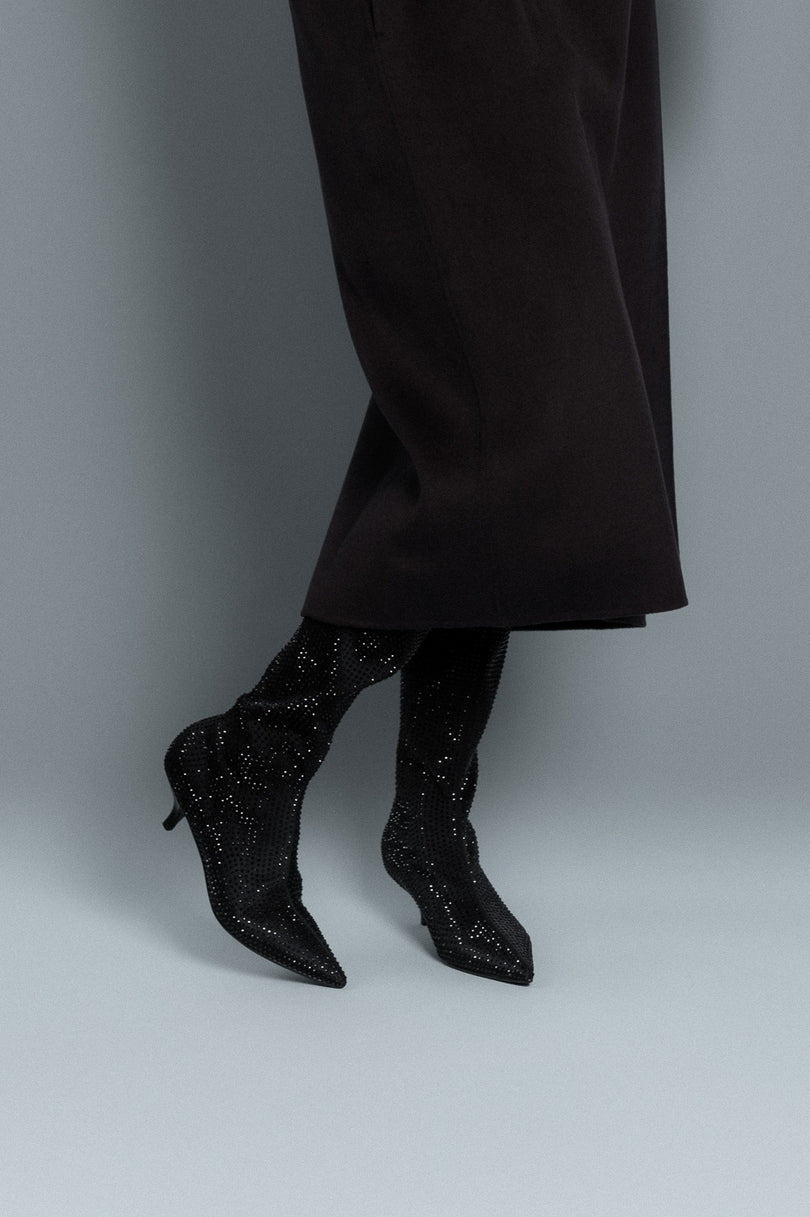 Black high-heeled shoes with sparkling details worn with black pants on a gray background