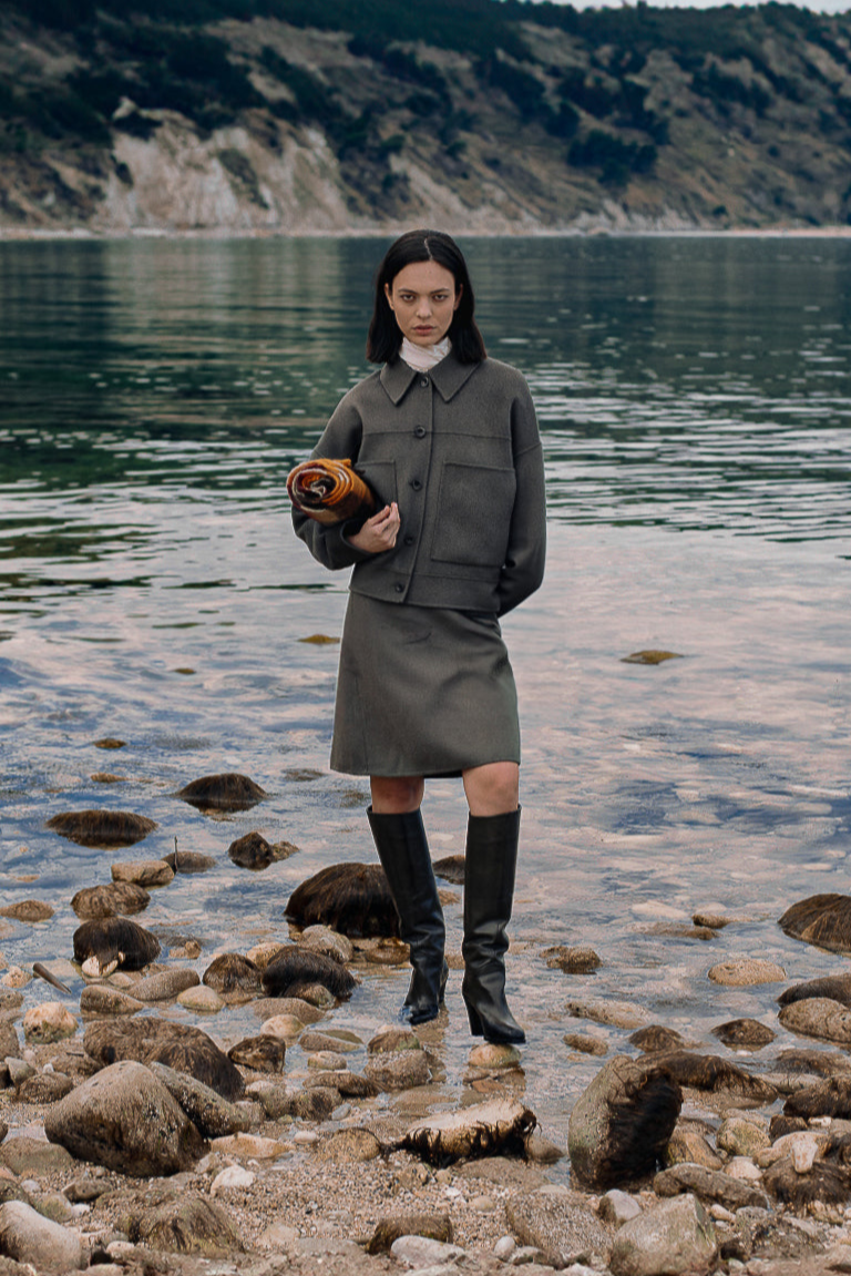 Person standing on a rocky shoreline holding a bag, with a scenic background of water and mountains.