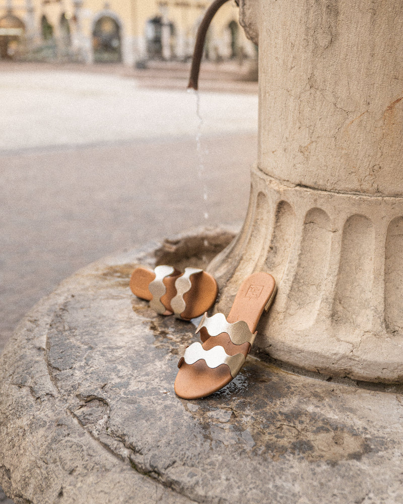 Pair of tan sandals with gold straps on a stone surface near a fountain.