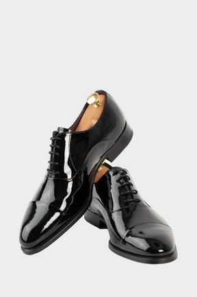 A pair of black patent leather lace-up oxford shoes with a cap toe, displayed on a light gray background.