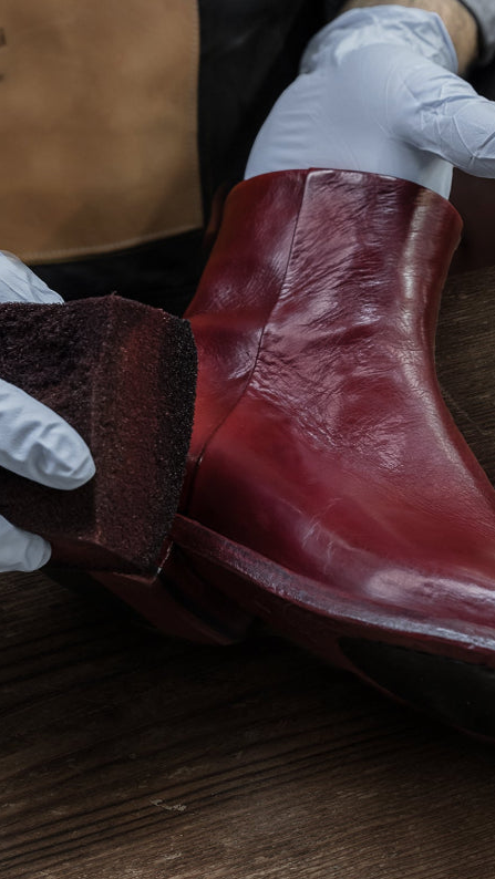 Person cleaning a red leather boot with a sponge on a wooden surface