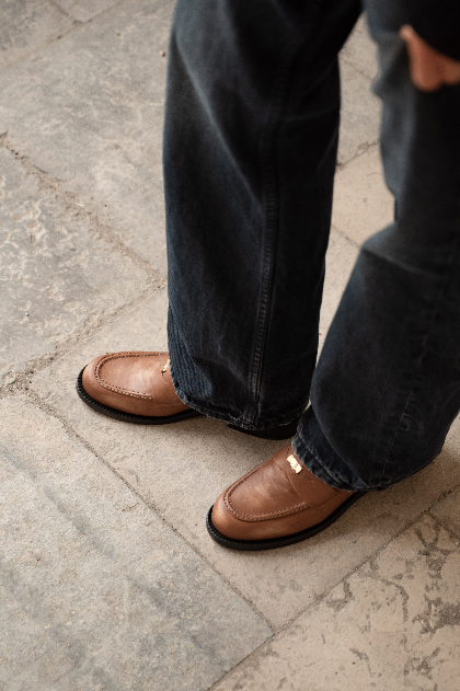 Brown leather shoes worn with dark blue jeans on a stone pavement.