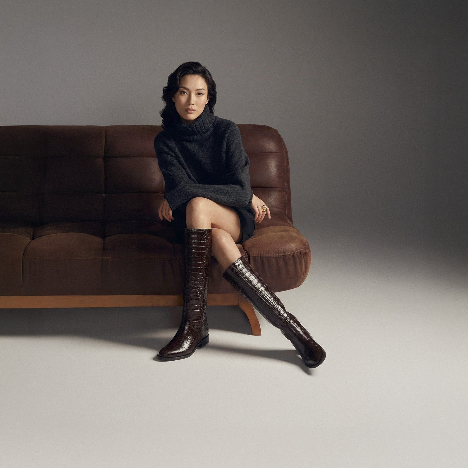 Woman sitting on a brown sofa wearing a black dress and knee-high boots against a gray background